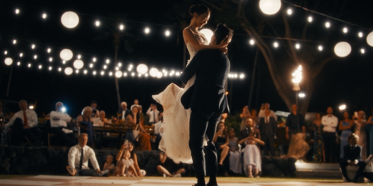 A groom lifts the bride beneath glowing reception lights as guests watch.