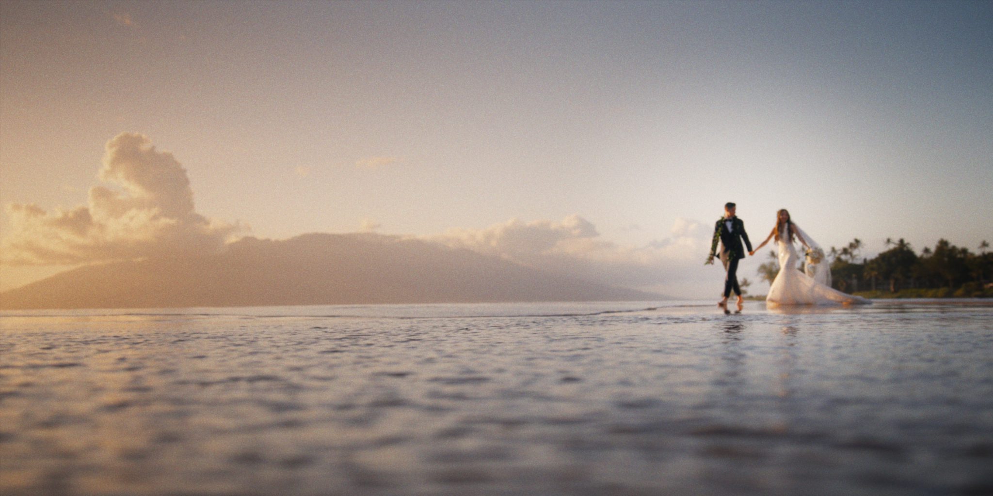 Lexi and Ryan walking at the ocean's edge under a wide sunset sky — Four Seasons Resort Maui.