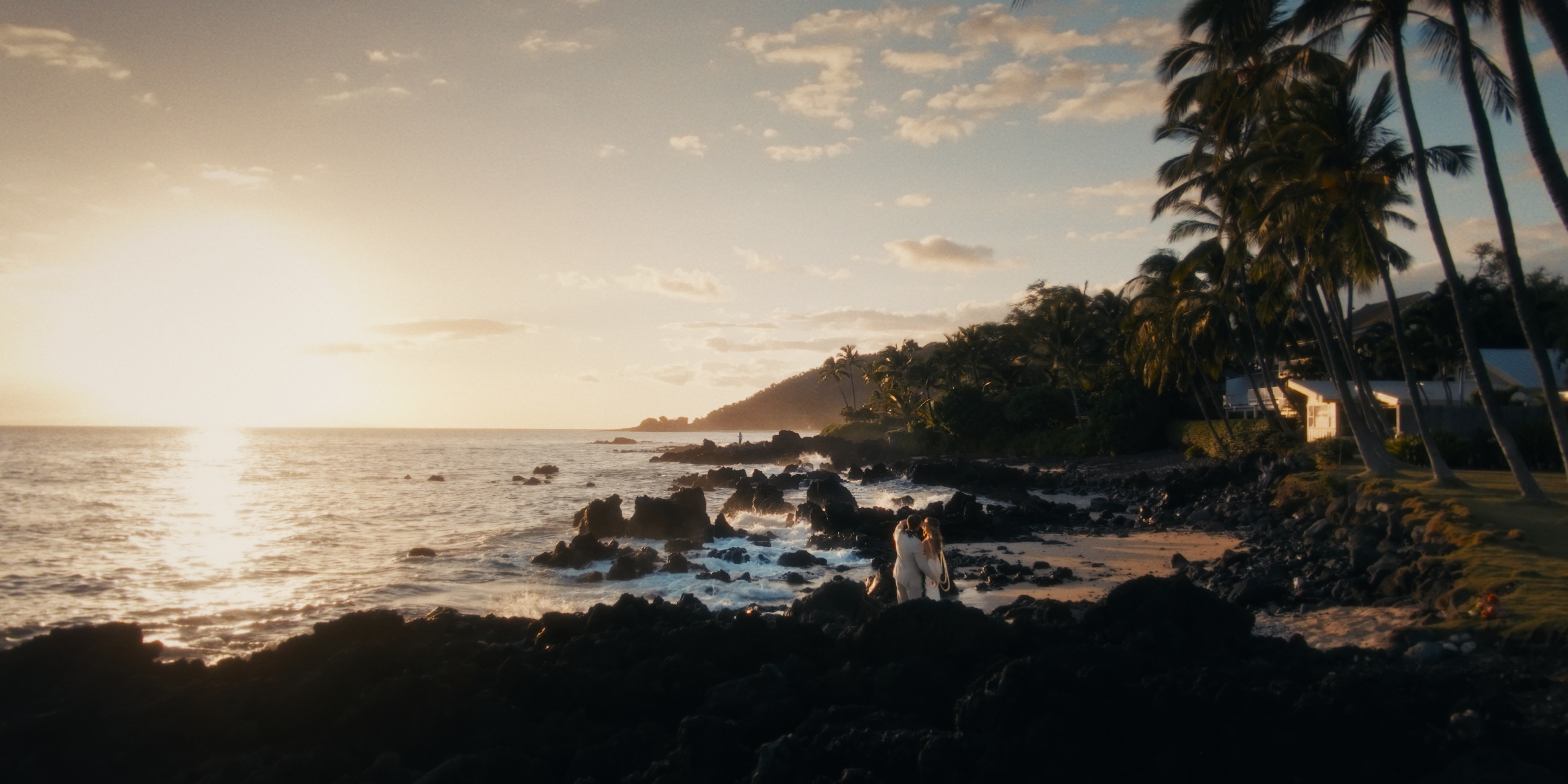 A couple embracing on the rocky Mākena shoreline at sunset, framed by the White Orchid Beach House palms